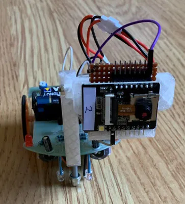 Robot pequeño de dos ruedas armado a mano, con placa de circuito, módulo de cámara y cables, sobre una mesa de madera