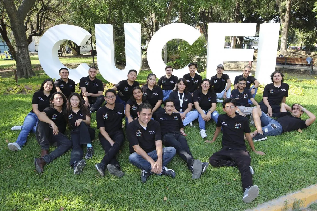 Grupo de estudiantes con playeras del equipo Mars Rover sentados sobre el pasto frente a las letras blancas de CUCEI