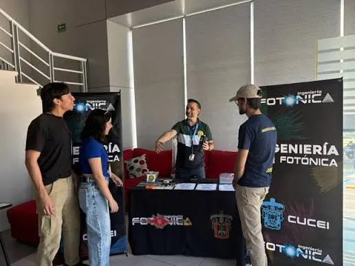 Cuatro personas conversando en el stand de Ingeniería Fotónica de CUCEI durante una feria de divulgación